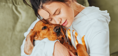 Lady lying on bed smiling with a brown dachshund puppy on her stomach with its tongue out